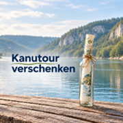 Hohe Glasflasche mit gerolltem Gutschein am Fluss und dem Schriftzug „Kanutour verschenken“.