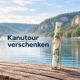Hohe Glasflasche mit gerolltem Gutschein am Fluss und dem Schriftzug „Kanutour verschenken“.
